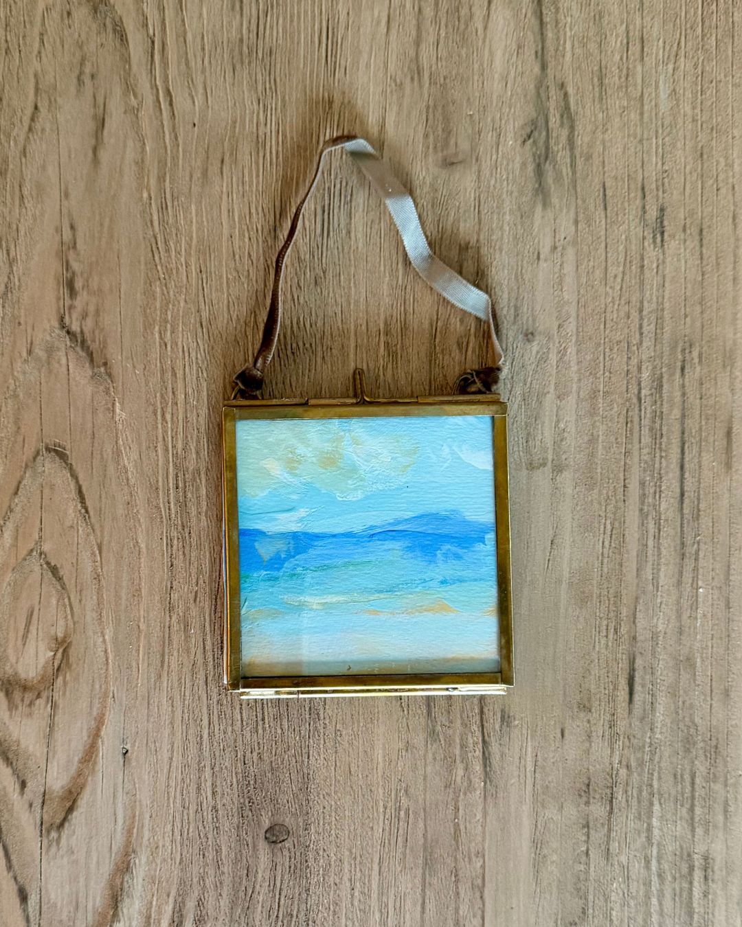 A small square framed landscape painting of a blue sea and sky, hanging from a white ribbon against a light brown wooden background.