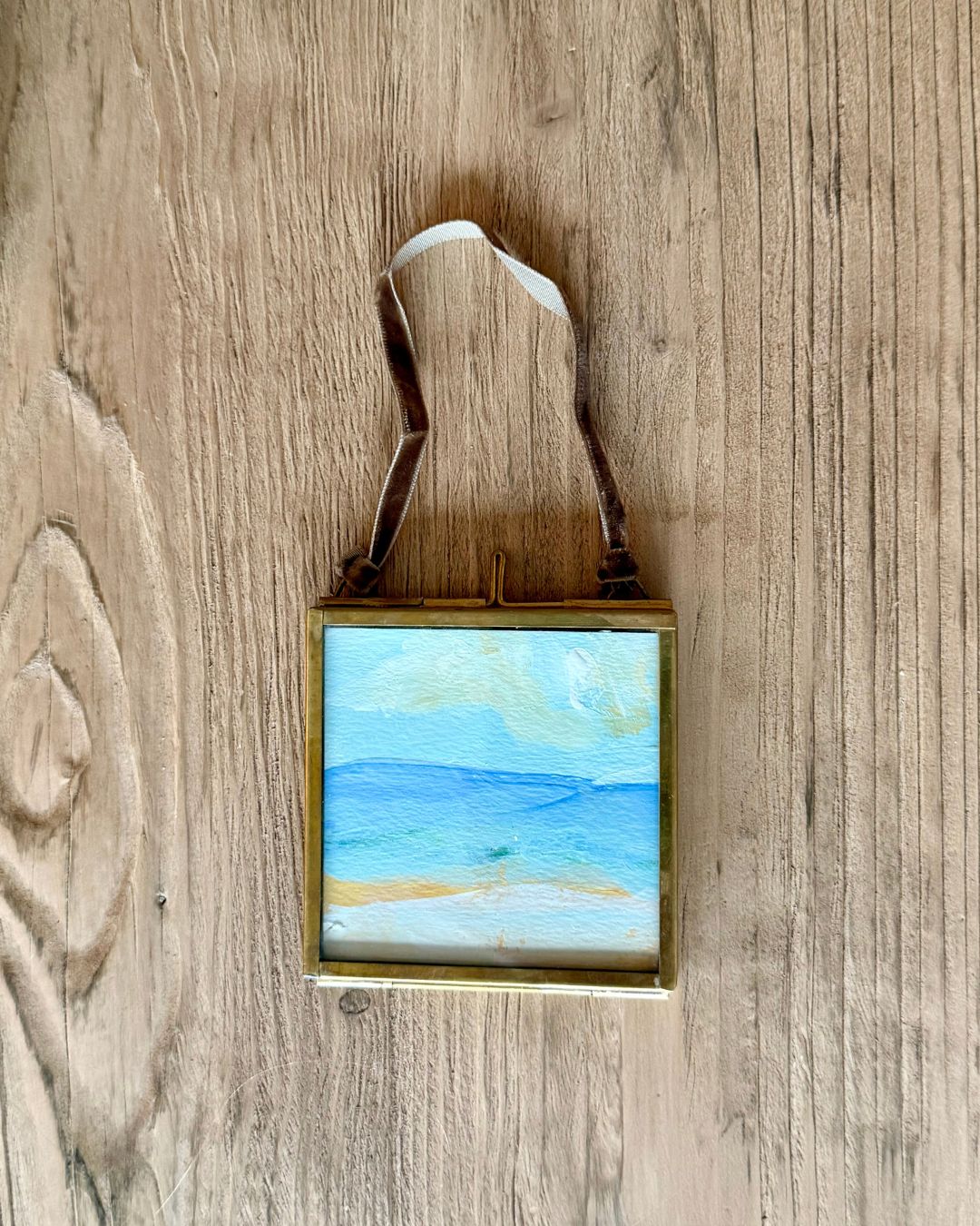 A small square framed painting of a beach landscape with blue water and sky, hanging on a rustic wooden wall by a brown ribbon.