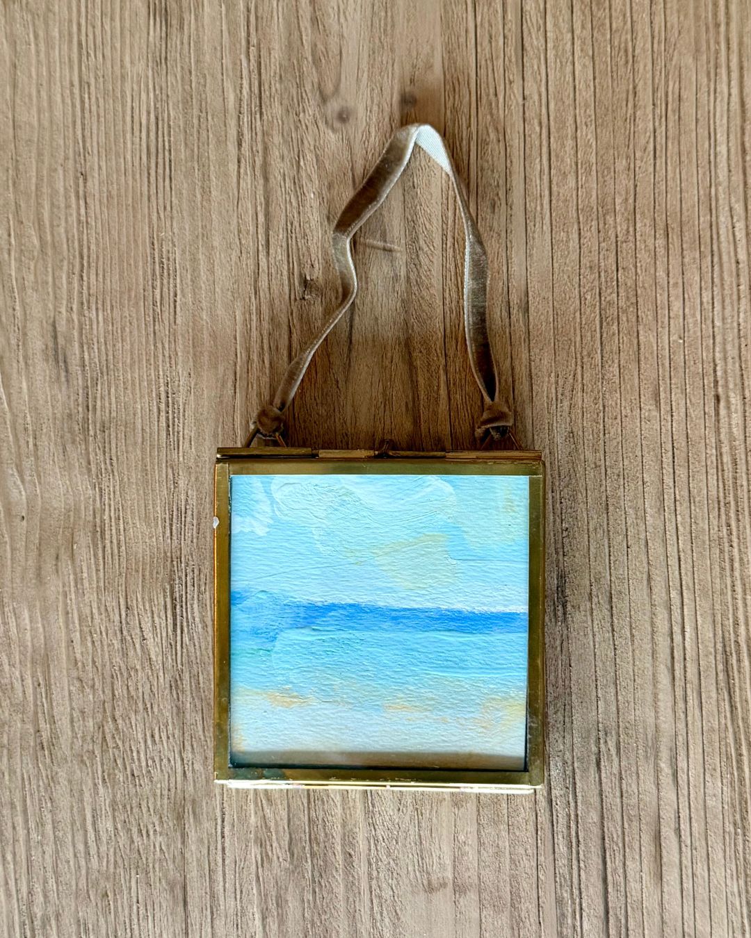 A small square framed painting of a blue ocean landscape with a gold-toned frame and a hanging ribbon, displayed on a wooden surface.
