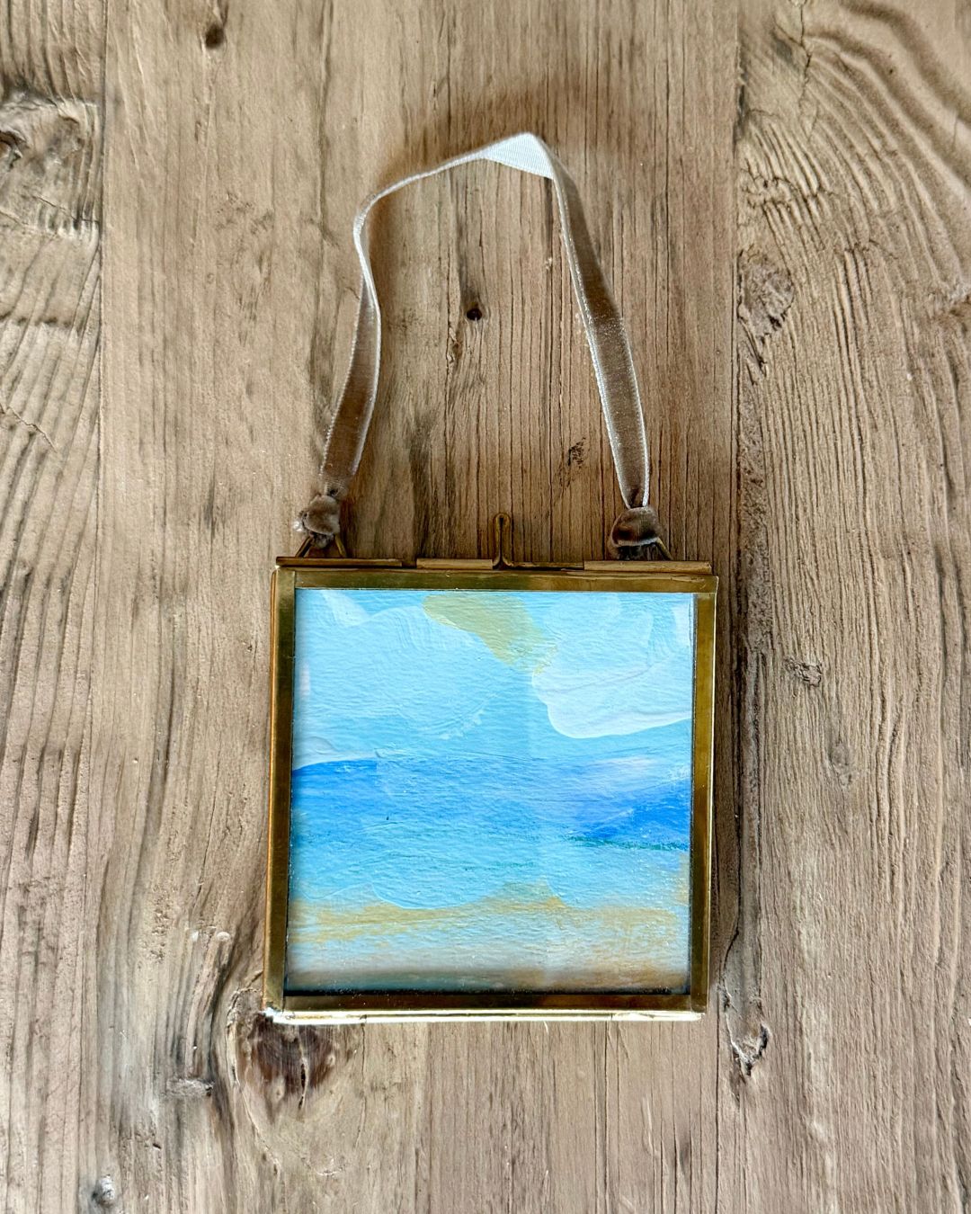 A small square framed painting with a blue sky and light blue ocean scene, hanging on a rustic wooden wall by a white ribbon.