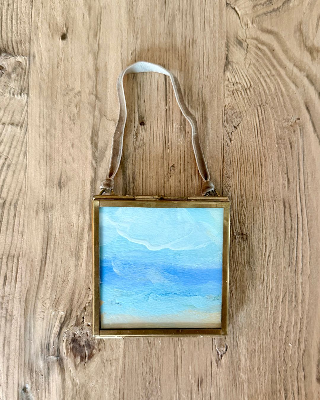 A small square framed art piece with a gold-toned frame and a white hanging ribbon, featuring an abstract blue and white landscape painting, displayed on a rustic wooden background.