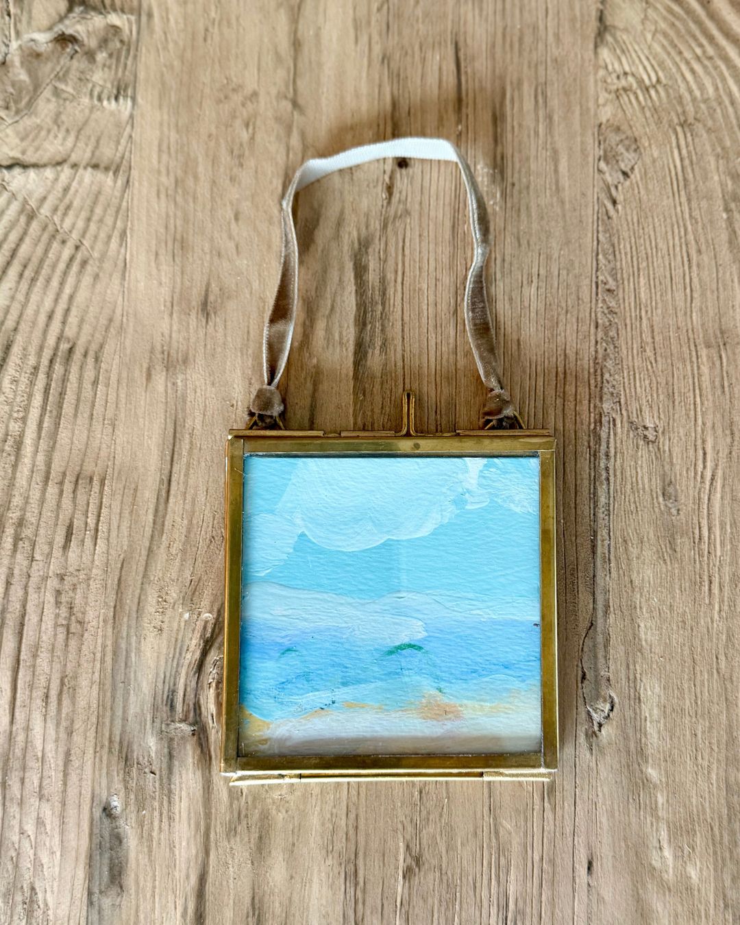 A square framed miniature painting of a beach scene with blue sky and ocean, hanging on a rustic wooden wall by a white ribbon.
