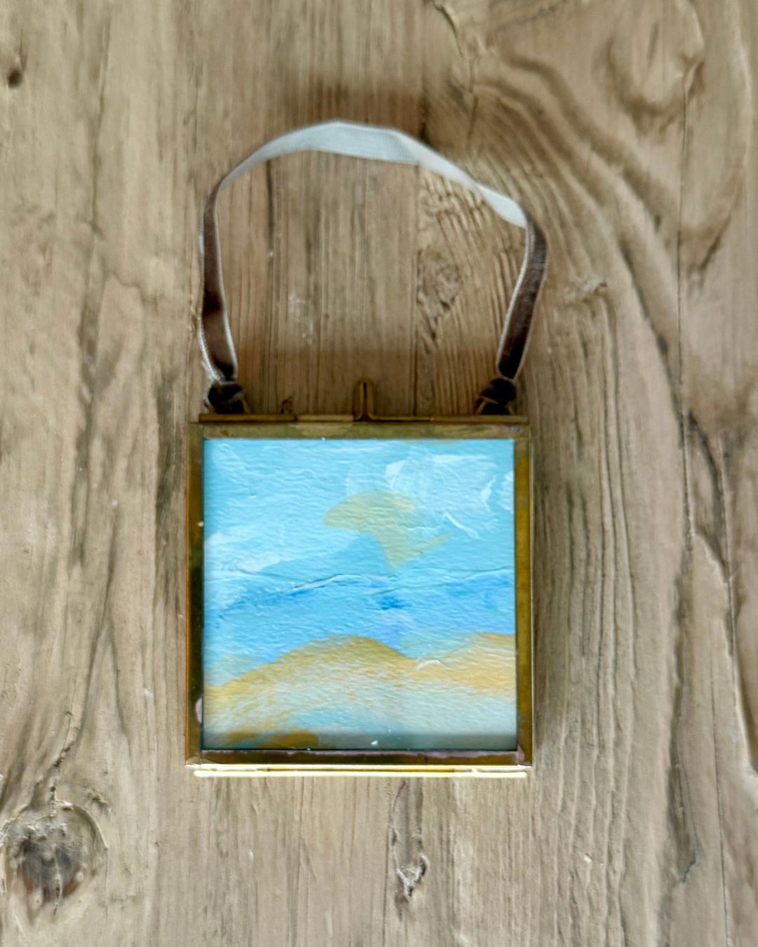 A square framed miniature landscape painting hanging on a rustic wooden surface. The artwork depicts a beach scene with blue sky and sandy yellow dunes, enclosed in a gold-toned frame with a dark brown leather loop at the top.