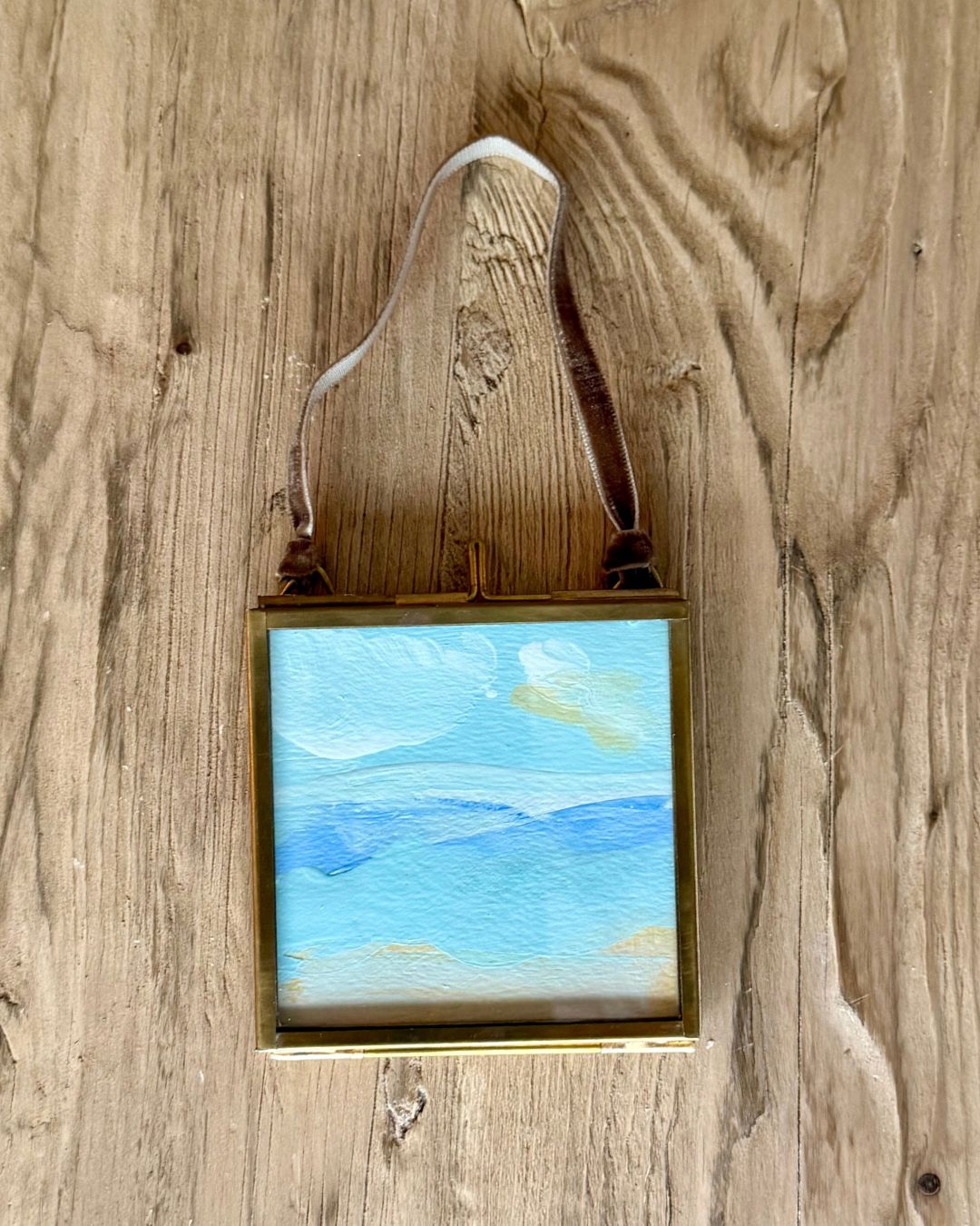 A small square framed landscape painting with blue and white sky and mountain scenes, hanging from a brown leather strap against a rustic wooden wall.