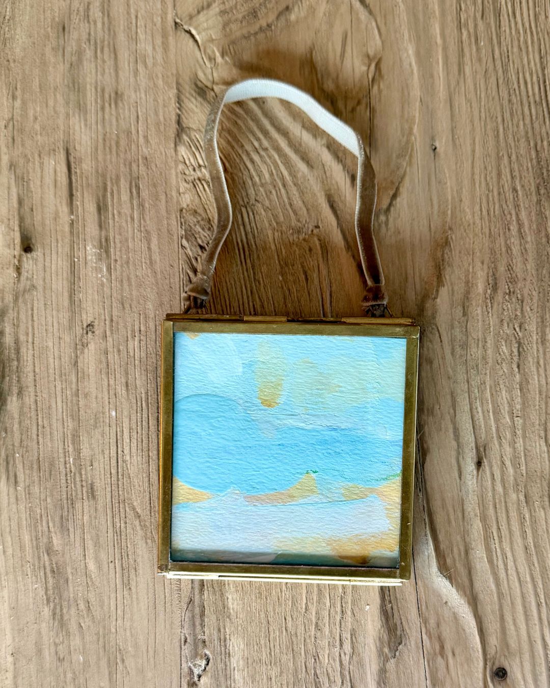 A square framed miniature landscape painting hanging on a rustic wooden wall. The painting depicts a beach scene with light blue water, white clouds, and golden yellow sunbeams, enclosed in a brass frame with a white hanging ribbon.