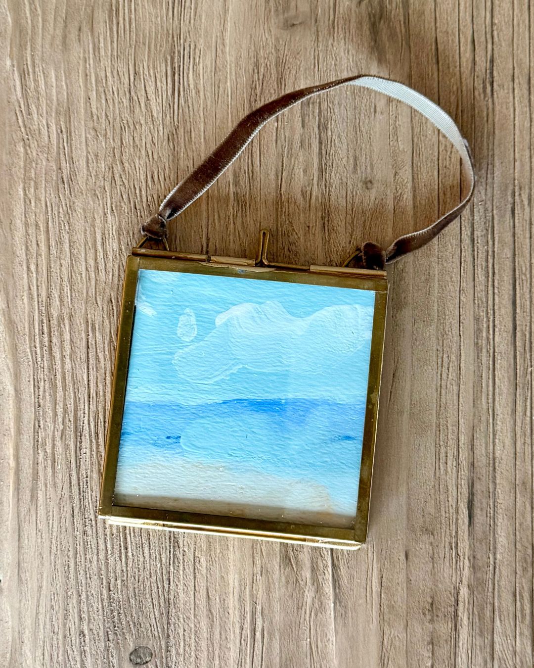 A square framed art print featuring a watercolor landscape of blue sky and white clouds, enclosed in a gold-toned frame with a brown leather strap, displayed on a wooden surface.