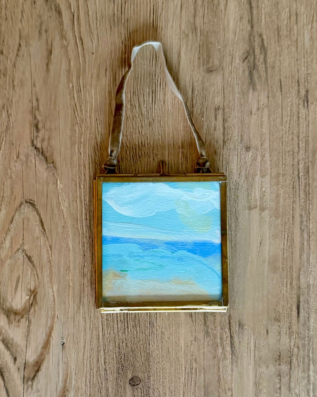 A small framed landscape painting of a beach scene with blue and white sky, hanging from a white ribbon against a rustic wooden background.